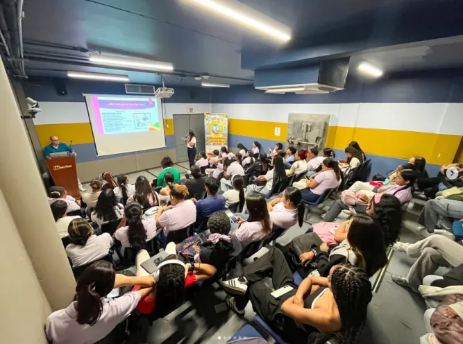 Estudiantes universitarios sentados en un auditorio oscuro asistiendo a una conferencia sobre igualdad de género frente a una pantalla de proyección.