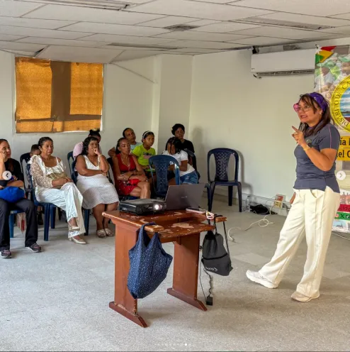 Facilitadora gesticulando frente a un atril y un proyector, mientras un grupo de mujeres asistentes la escucha sentadas en sillas plásticas dentro de un salón.