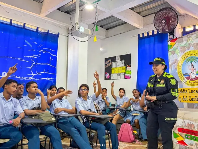 Oficial de la Policía de Infancia y Adolescencia impartiendo una charla de prevención a estudiantes de secundaria que levantan la mano en un salón de clases.