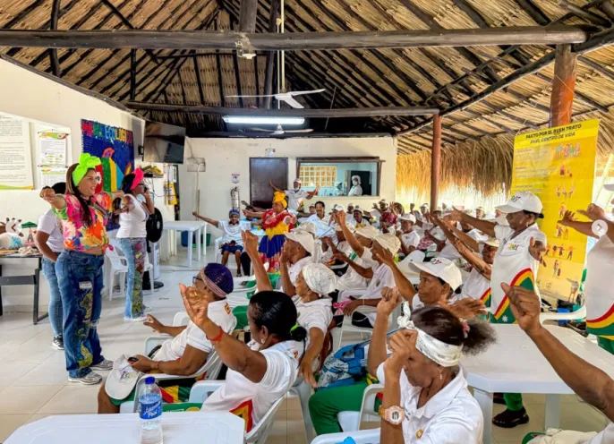 Facilitadora guiando dinámicas de recreación activa con un grupo numeroso de adultos mayores sentados en mesas dentro de un salón comunal techado en Barú.