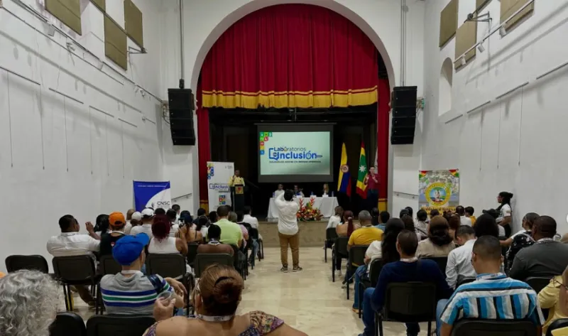 Asistentes sentados en un auditorio frente a un escenario con una pantalla que proyecta el evento "Laboratorios de Inclusión".