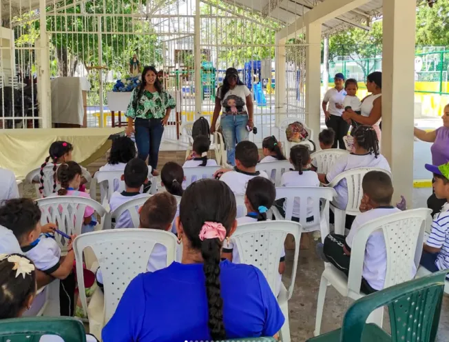 En la imagen se observa un grupo de niños y adultos sentados en sillas plásticas participando en una charla educativa al aire libre, con dos mujeres de pie al frente dirigiéndose a los asistentes.