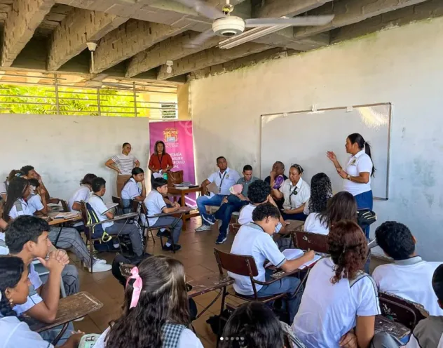 En el salón de clases, un grupo de estudiantes uniformados atiende atentamente una charla impartida por una mujer de pie junto al tablero.