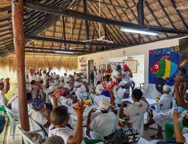 Un grupo numeroso de personas mayores participa alegremente en una actividad cultural en un espacio techado con techo de palma, mientras algunas personas al frente guían la dinámica.