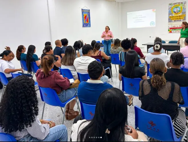 En la imagen se aprecia a un numeroso grupo de asistentes sentados en sillas azules dentro de un auditorio, prestando atención a una mujer que imparte una capacitación frente a una pantalla de proyección.