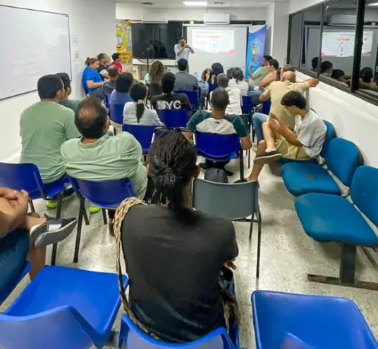 En la imagen se observa un auditorio lleno de asistentes sentados en sillas azules, mientras una persona de pie al fondo imparte una presentación frente a una pantalla proyectada.