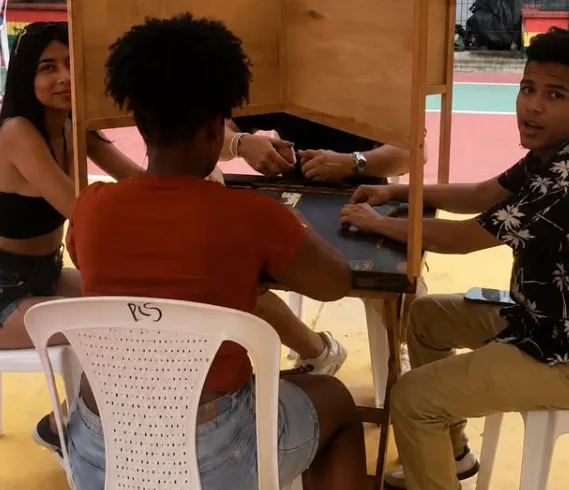 Un grupo de jóvenes se encuentra sentado alrededor de una mesa compartiendo y participando en un juego de dominó al aire libre.