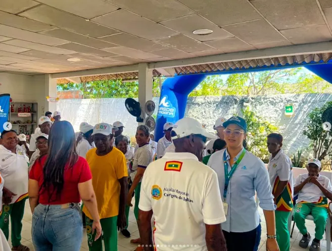 Fotografía de una jornada ambiental en el Centro de Vida Zapatero con adultos mayores y personal institucional. En primer plano, personas de pie interactuando en un espacio iluminado, incluyendo a una mujer con camisa azul claro y gorra, y hombres con camisetas blancas y amarillas con detalles institucionales. Al fondo se aprecia una lona azul y un espacio abierto con vegetación.