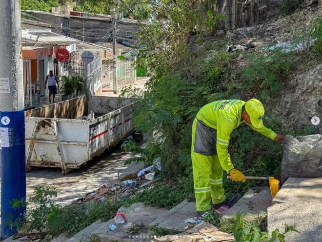 Fotografía tomada al aire libre en una calle urbana en pendiente. En primer plano, un operario con uniforme reflectivo verde y gorra amarilla se encuentra agachado sobre unas escaleras de concreto, barriendo y recogiendo residuos junto a una bolsa negra. A la izquierda, un contenedor metálico grande para escombros o basura está ubicado al lado de una calle angosta con viviendas y vegetación de fondo.