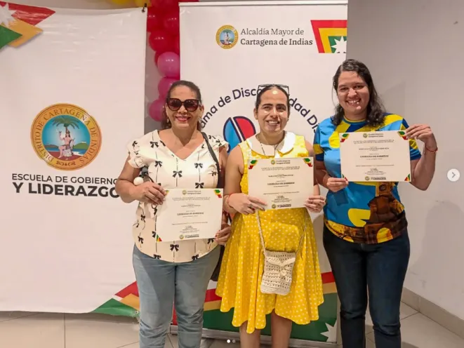 Fotografía en un espacio interior decorado con pancartas institucionales donde tres mujeres sonríen sosteniendo sus certificados de culminación del proceso de formación "Liderazgo Sin Barreras".