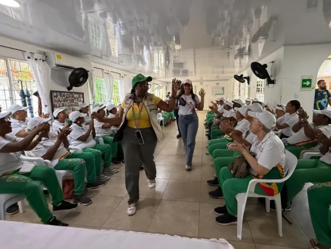 En la imagen se observa a un grupo de personas mayores sentadas en un salón aplaudiendo mientras dos mujeres caminan por el pasillo central durante la jornada ambiental.