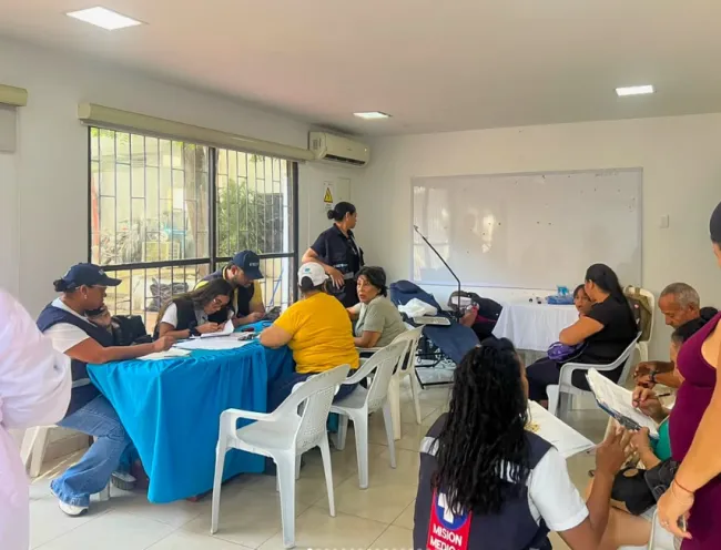 Fotografía en un espacio interior iluminado donde personal de salud y de atención ciudadana atienden a varias personas sentadas alrededor de mesas y escritorios organizados para una jornada de servicios integrales.