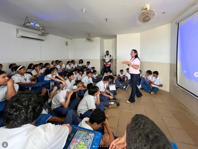 Fotografía tomada en un salón de clases donde una conferencista de pie dirige una charla a un grupo numeroso de estudiantes sentados en pupitres y en el piso frente a una pantalla de proyección.   