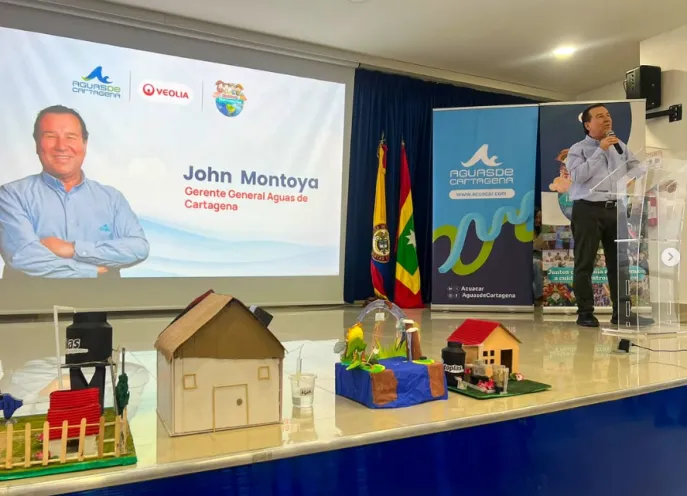 Fotografía en un auditorio donde un hombre habla ante un atril, con una pantalla gigante al fondo que muestra la imagen de un directivo y los logos de Aguas de Cartagena y Veolia. En una mesa delantera se exhiben varias maquetas artesanales de casas y proyectos escolares.   