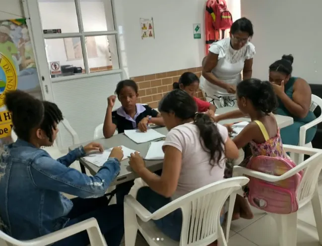 En la imagen se observa a un grupo de niños y adolescentes sentados alrededor de una mesa participando en una dinámica de formación y desarrollo personal, acompañados por talleristas.