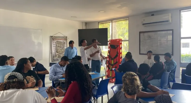 En la imagen se aprecia a un instructor señalando hacia una camilla de emergencia frente a un salón de clases lleno de estudiantes atentos.