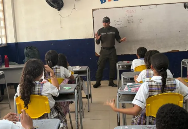 En la imagen se observa a un oficial de policía impartiendo una charla de prevención a los estudiantes en un salón de clases.