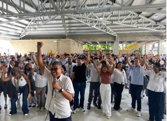Fotografía en un amplio espacio cubierto donde un grupo numeroso de jóvenes estudiantes y coordinadores celebran con los brazos en alto, liderados por un hombre con micrófono en primer plano.   