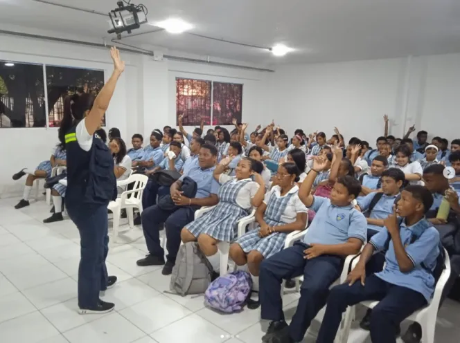 Fotografía en un salón de clases iluminado donde una instructora de pie levanta el brazo izquierdo frente a decenas de estudiantes con uniforme escolar sentados en sillas plásticas, quienes participan activamente en una charla de prevención.
