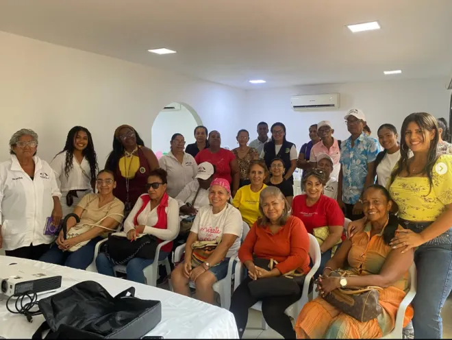 Fotografía grupal en un salón interior iluminado donde un grupo numeroso de líderes comunitarios, en su mayoría mujeres sentadas y de pie, posan sonrientes para la cámara durante una jornada institucional.   