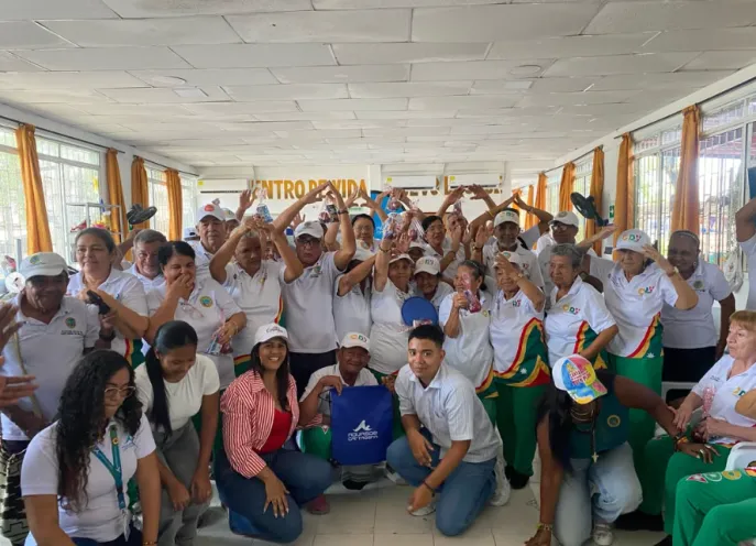 Fotografía en un salón interior iluminado donde un grupo numeroso de adultos mayores y personal institucional posan alegres con los brazos en alto, celebrando una jornada lúdico-ambiental en el Centro de Vida del Nuevo Bosque.