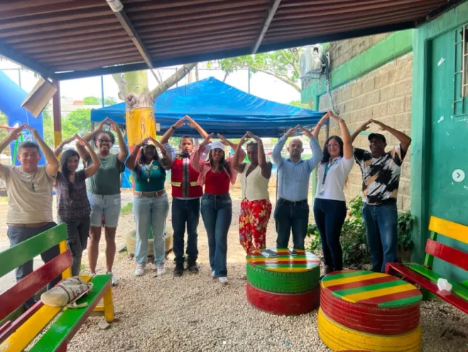 Fotografía al aire libre bajo un techo de zinc donde un grupo de líderes comunitarios y representantes de la Alcaldía Local 1 posan sonrientes haciendo gestos con las manos sobre la cabeza, junto a mesas hechas con llantas recicladas de colores.