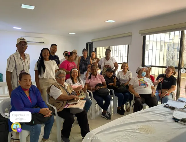 Fotografía en un salón interior iluminado donde un grupo de líderes comunales de la Unidad Comunera 10 se encuentra reunido, algunos sentados en sillas y otros de pie, participando en un taller de sensibilización ambiental.   