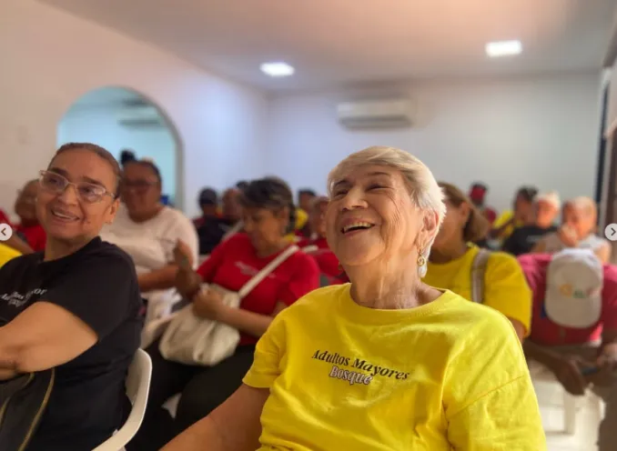 Fotografía en un espacio interior iluminado donde adultos mayores participan sonrientes en una jornada lúdico-ambiental, destacando en primer plano una mujer con una camisa amarilla que lleva la inscripción "Adultos Mayores Bosque".
