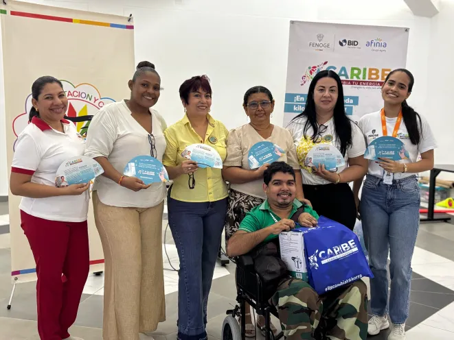 Grupo de mujeres y un joven en silla de ruedas posan sonrientes sosteniendo abanicos informativos y una bolsa azul institucional de la estrategia "Caribe Cambia Tu Energía", frente a pancartas de FENOGE, BID y Afinia.