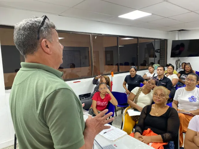 Un instructor de espaldas dirige una clase a un grupo de ciudadanos sentados en un aula, quienes asisten atentos a la capacitacion de atencion al cliente.