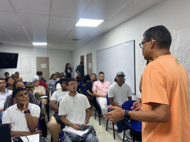 Un instructor con camisa naranja de espaldas gesticula mientras dirige una charla a un grupo de personas y jovenes sentados atentamente en un aula de capacitacion.